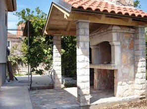 A stone-built outdoor oven structure with a tiled roof, surrounded by a stone-paved area and greenery.