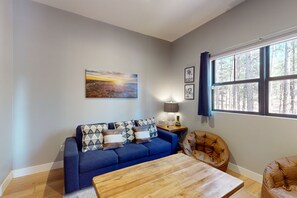 A living room with a blue sofa, a wooden coffee table, and a large window with a view of trees. - Updated luxury home with ensuite bedrooms, fireplace, & large deck w/ firepit (Flagstaff)