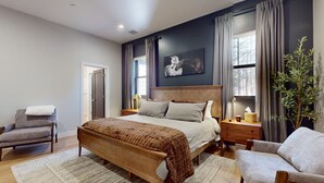 A bedroom with a large bed, a chair, a nightstand, a lamp, a window with curtains, and a painting on the wall. - Updated luxury home with ensuite bedrooms, fireplace, & large deck w/ firepit (Flagstaff)