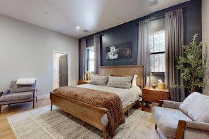 A bedroom with a large bed, a chair, a nightstand, a lamp, a window with curtains, and a painting on the wall. - Updated luxury home with ensuite bedrooms, fireplace, & large deck w/ firepit (Flagstaff)