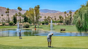 A golf course with a pond, palm trees, and mountains in the background. - Bright and cheery home close to Coachella with pool & tennis on-site (Palm Desert)