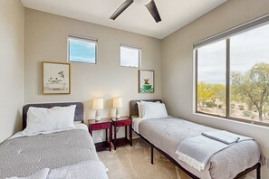 Ein Schlafzimmer mit zwei Einzelbetten, einem Deckenventilator, zwei Nachttischen mit Lampen, gerahmten Bildern an den Wänden und einem großen Fenster mit Blick auf Bäume.