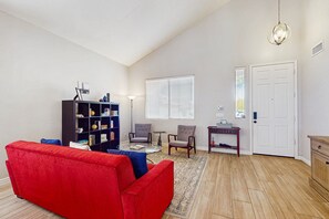 Ein Wohnzimmer mit einem roten Sofa, einem dunkelblauen Bücherregal, zwei Sesseln, einem kleinen runden Tisch und einem großen Fenster mit Jalousien.