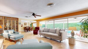 A spacious living room with a beige sofa, a blue armchair, and a red chair. There is a glass coffee table and a ceiling fan. - Eclectic  mid-century, dog-friendly home with private pool, yard & gas grill (Palm Springs)