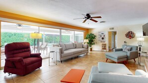 A spacious living room with a red recliner, grey sofa, and a small orange table. There are ceiling fans, a large window with a view of greenery, and a plant in the corner. - Eclectic  mid-century, dog-friendly home with private pool, yard & gas grill (Palm Springs)
