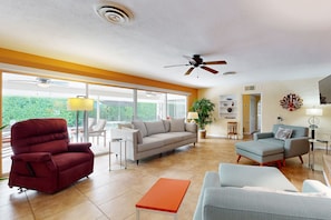 Ein geräumiges Wohnzimmer mit einem roten Liegesessel, einem grauen Sofa und einem kleinen orangefarbenen Tisch. Es gibt Deckenventilatoren, ein großes Fenster mit Blick auf Grünflächen und eine Pflanze in der Ecke.