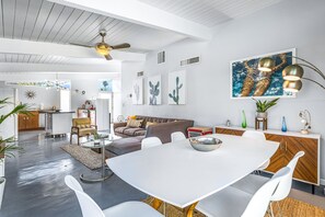 A modern living room with a dining area, featuring a white table, white chairs, a brown sofa, and a ceiling fan. - Iconic, bright, & dog-friendly house w/private pool, spa, pool table & grill (Palm Springs)