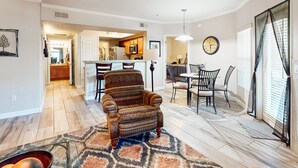 Un salón amplio con un sillón estampado, una mesa de comedor con sillas, una cocina al fondo y un ventanal con persianas.