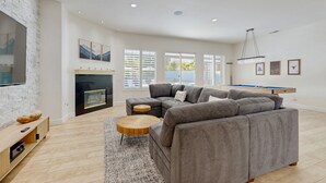 A modern living room with a sectional sofa, a fireplace, a pool table, and large windows.