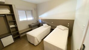 Una habitación pequeña y limpia con dos camas individuales, un escritorio, una silla, un armario y un refrigerador pequeño.