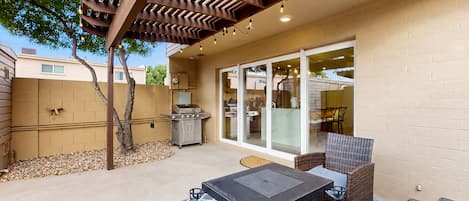 Een terras buiten met een barbecue, rieten meubilair en een pergola met lichtslingers.