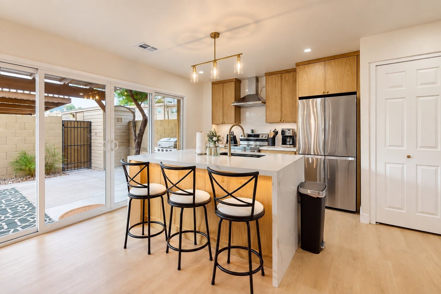 Una cocina moderna con una isla central, taburetes de bar, electrodomésticos de acero inoxidable y un patio al que se accede por una puerta corredera de vidrio.