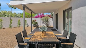 Een terras buiten met een tafel en stoelen, een overdekt gedeelte en uitzicht op een tuin met roze bloemen.