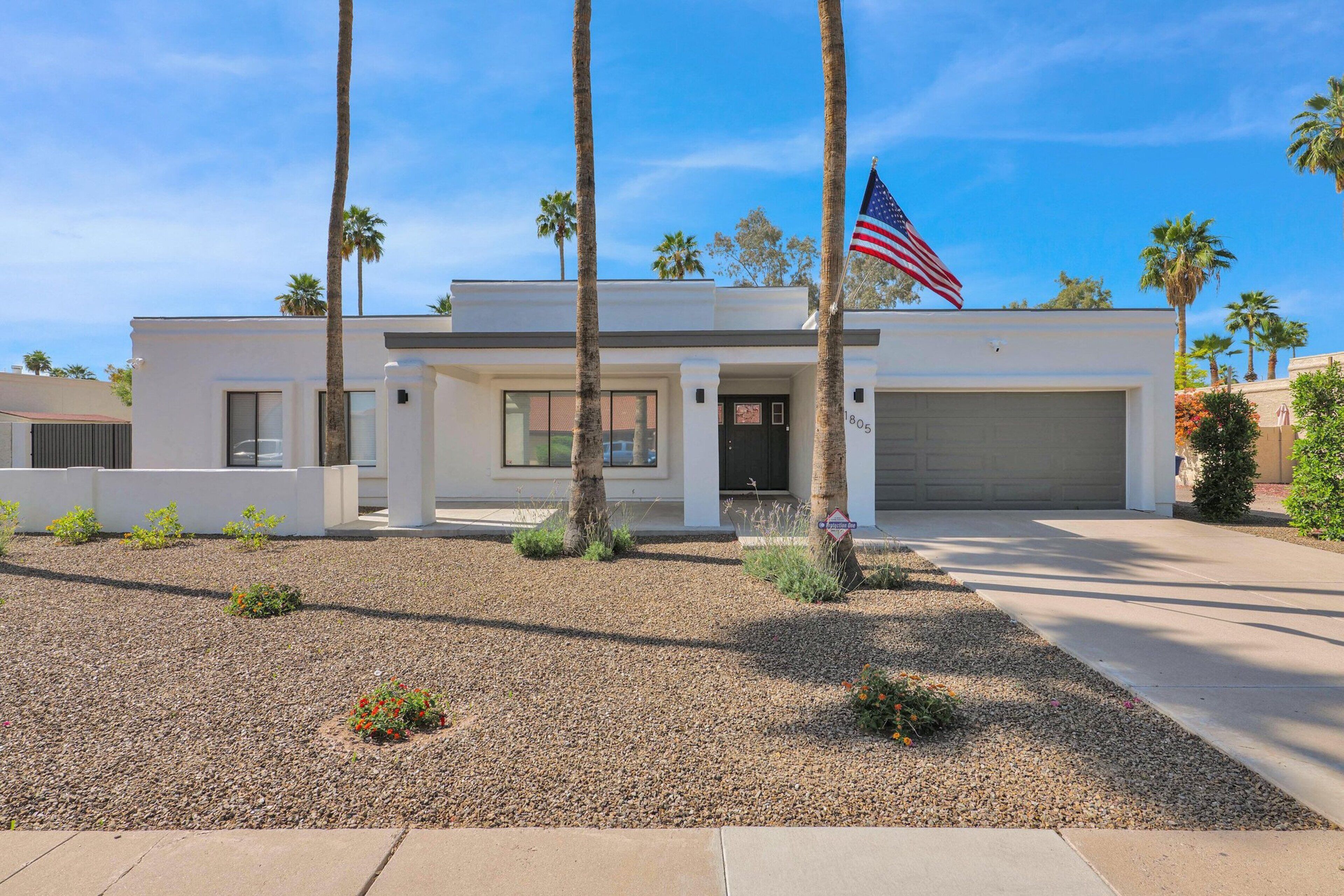 A modern two-story house with a garage, palm trees, and an American flag.