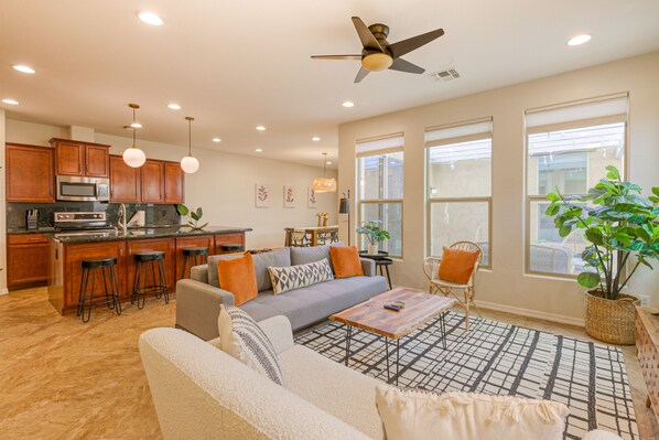 A modern kitchen with wooden cabinets, a microwave, and bar stools. There is a gray sofa with orange cushions, a coffee table, and a ceiling fan.