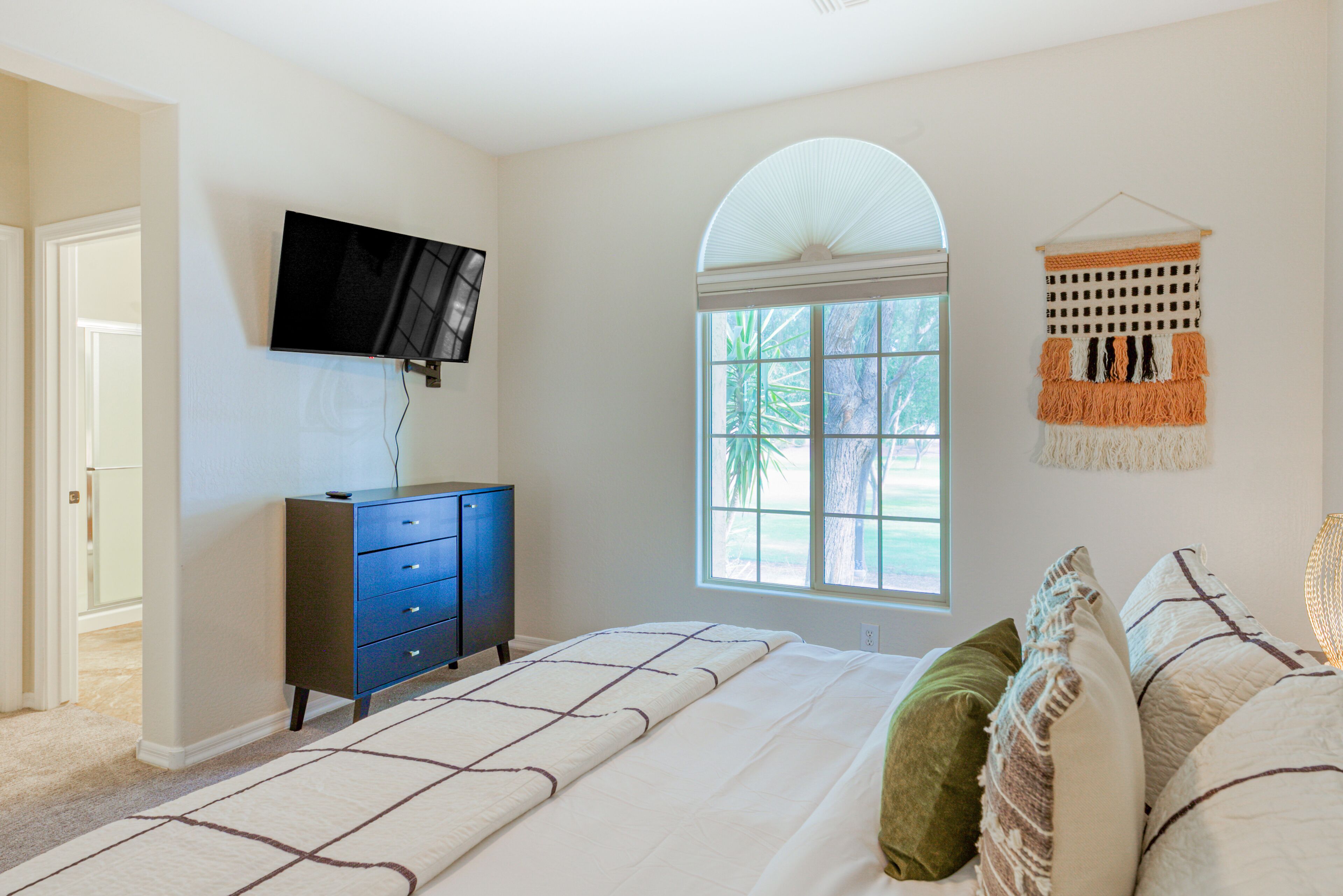 A bedroom with a bed, a dresser, a TV, a window with a view, and a macrame wall hanging.