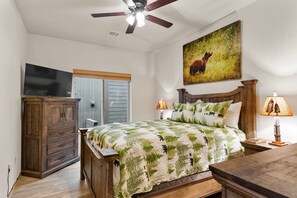 Ein Schlafzimmer mit Bett, Holz-Kopfteil, Deckenventilator, Fernseher an der Wand, einem Gemälde und einem Nachttisch mit Lampe.