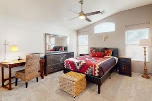 Un dormitorio con cama, escritorio, silla, pequeño reposapiés, cómoda, espejo y ventilador de techo.