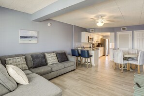 Una sala de estar con un sofá modular gris, un comedor con sillas a rayas y una cocina al fondo.