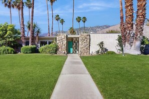 A pathway leading to a building with a stone facade and a green door, surrounded by palm trees and a clear sky. - Stylish Dog-Friendly Retreat W/ High-Speed WiFi, Pool & Spa, & Pool Table (Palm Springs)