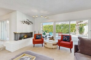 A modern living room with a fireplace, two red armchairs, a grey sofa, and a view of a pool and greenery outside. - Stylish Dog-Friendly Retreat W/ High-Speed WiFi, Pool & Spa, & Pool Table (Palm Springs)