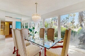A dining area with a glass table and chairs, a chandelier, and a view of palm trees. - Stylish Dog-Friendly Retreat W/ High-Speed WiFi, Pool & Spa, & Pool Table (Palm Springs)