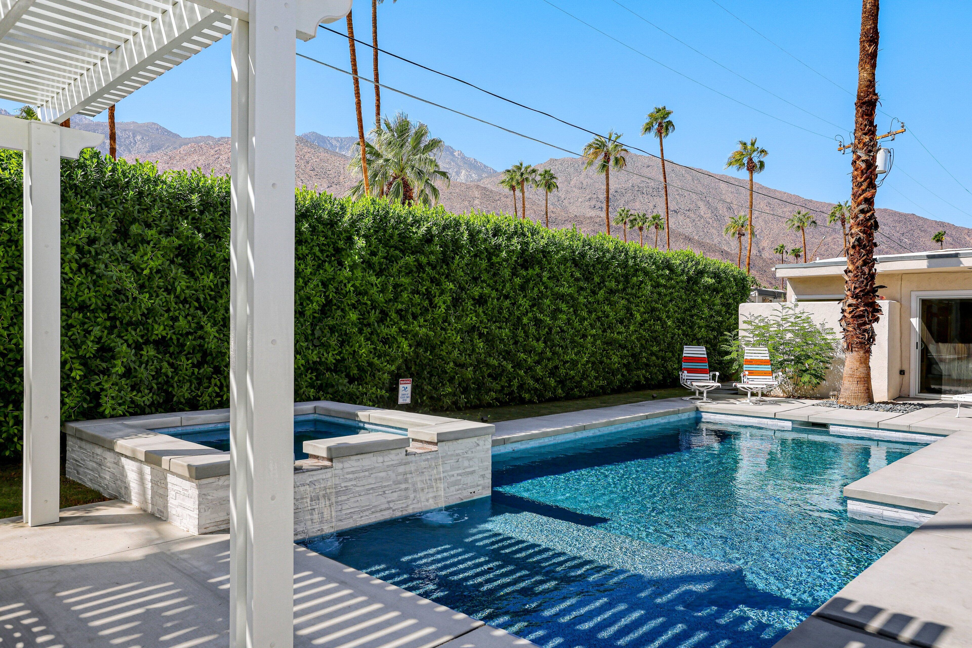 A swimming pool with a white pergola, palm trees, and a mountain backdrop.