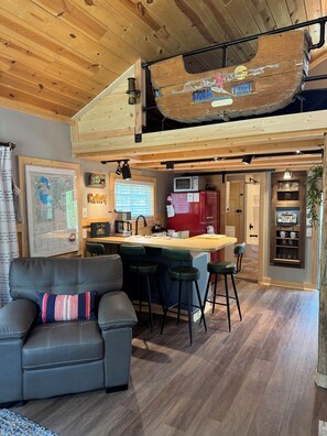 A loft-style living space with a wooden ceiling, a bar area, a kitchen, and a seating area with a couch and stools.