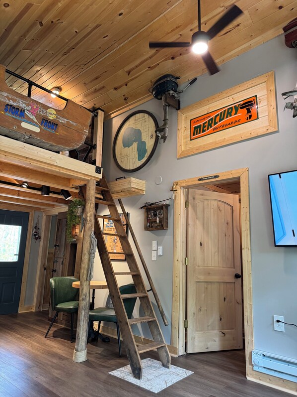 A loft-style room with a wooden ceiling, a loft bed, a wooden ladder, a green chair, a wooden door, and a wall-mounted TV.