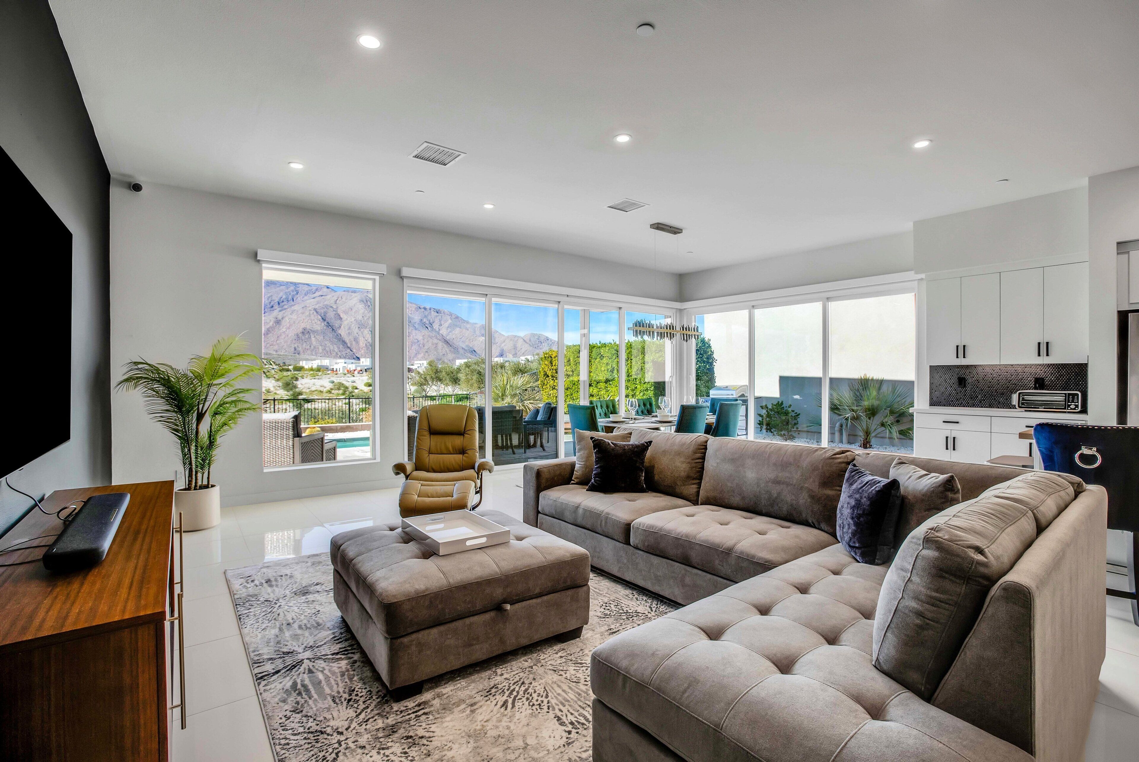 A modern living room with a large sectional sofa, a coffee table, and a TV on a wooden stand. There is a large window with a view of mountains and a pool.