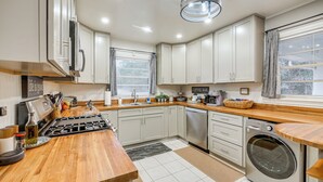 Een moderne keuken met witte kasten, een houten aanrecht, een ingebouwde kookplaat, een wasmachine en een bord met daarop ‘koffiebar’.