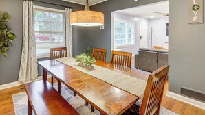 Een eethoek met een houten tafel, stoelen, een hanglamp en uitzicht op een woonkamer door een geopende deur.