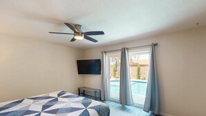 Un dormitorio con cama, ventilador de techo, televisor y vista a una piscina a través de amplios ventanales.