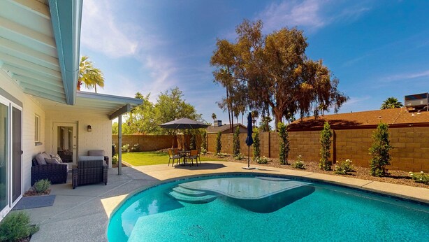 Una piscina con borde curvo, rodeada por un patio con mobiliario exterior y una zona de estar cubierta.