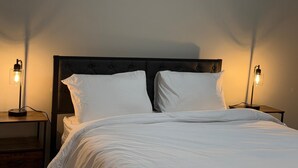 Ein ordentlich bezogenes Bett mit weißen Leinen, an beiden Seiten mit Nachttischen und Lampen, in einem Raum mit einem dunkel gepolsterten Kopfteil.