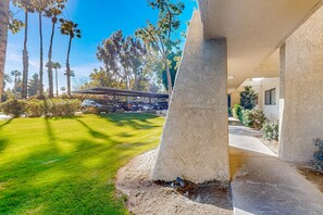 A modern building with a covered outdoor area, a parking lot, and a well-maintained lawn with tall palm trees. - Snowbird-friendly retreat with pool, hot tub, tennis, putting green, & patio (Palm Springs)