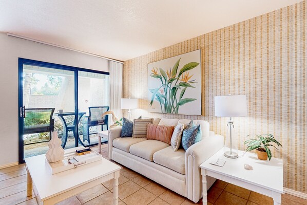 A living room with a sofa, coffee table, and a potted plant. There is a window with a view of a garden and a door leading outside. - Snowbird-friendly retreat with pool, hot tub, tennis, putting green, & patio (Palm Springs)
