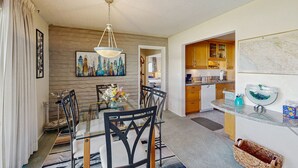 A dining area with a glass table and chairs, a hanging light fixture, a painting, and a kitchen in the background. - Sunny Palm Springs Retreat with Shared Pool, Hot Tub & Prime Location! (Palm Springs)