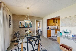 A dining area with a glass table and chairs, a hanging light fixture, a painting, and a kitchen in the background. - Sunny Palm Springs Retreat with Shared Pool, Hot Tub & Prime Location! (Palm Springs)