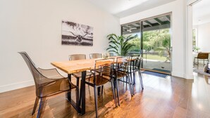 A modern dining area with a wooden table and clear plastic chairs, a large window, and a door leading to a patio. - Stylish home with pool & spa, ping-pong table, WiFi & fireplace - great location (Paradise Valley)