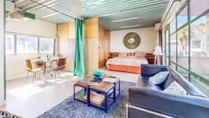 Una habitación de hotel moderna con zona de comedor, sofá, cama con ropa de cama estampada y ventanas grandes que ofrecen vistas.