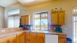A kitchen with wooden cabinets, a dishwasher, and a window with a view of palm trees. - Airy home with private heated pool, firepit, grill, enclosed backyard, cornhole (Goodyear)