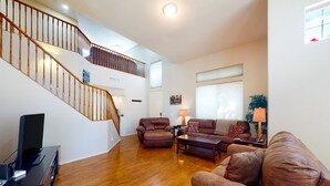 A spacious living room with hardwood flooring, a brown leather sofa, a coffee table, and a TV on a stand. A staircase with wooden railings leads to an upper level. - Airy home with private heated pool, firepit, grill, enclosed backyard, cornhole (Goodyear)