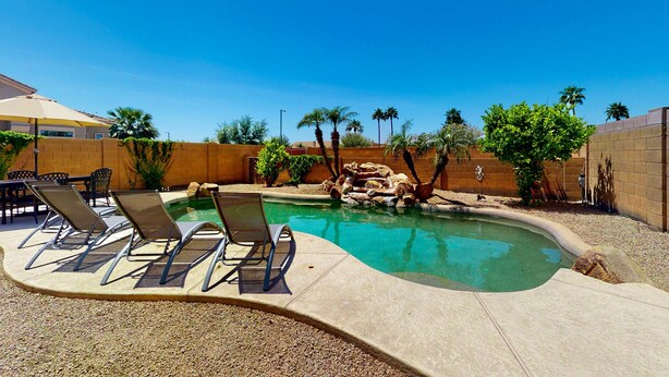 A swimming pool with lounge chairs, palm trees, and a brick wall. - Airy home with private heated pool, firepit, grill, enclosed backyard, cornhole (Goodyear)