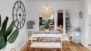 Un coin repas avec une table blanche, un banc et des chaises. On y trouve une grande horloge murale, un luminaire suspendu et une plante.
