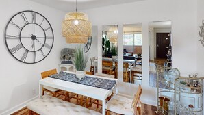 Un coin repas avec une table blanche, des chaises en bois et une grande horloge murale. Un luminaire suspendu en rotin se trouve au-dessus de la table.