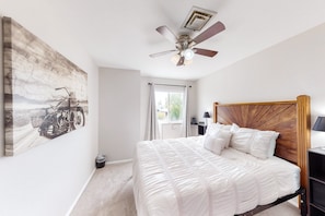 Une chambre à coucher comprenant un lit, un ventilateur de plafond, une fenêtre avec des rideaux et une œuvre d’art accrochée au mur.