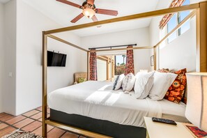 Ein Bett mit Baldachin, ein an der Wand befestigter Fernseher, ein Deckenventilator und Blick ins Freie durch eine offene Tür.