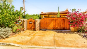 A gated entrance with a wooden fence, a concrete driveway, and lush greenery including pink flowers and a palm tree. - Mountain-view home with private pool & spa, near trails - 3BR #220080 (La Quinta)