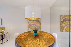 Uma área de refeições moderna com uma mesa redonda de madeira, cadeiras brancas, uma planta em vaso central e uma grande luminária pendente com textura.